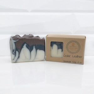 May include: A handmade bar of soap with a marbled design in shades of brown, gray, and white. The soap is next to a small cardboard box with a window showing a smaller soap sample. The box reads "Cedar Leather" and "Handmade Soap 4 oz."