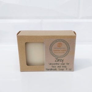 Op de afbeelding: Een staafje ongeparfumeerde handgemaakte zeep in een bruine kartonnen doos. De zeep is gelabeld "Zincy" en is voor gezicht en lichaam. Het label vermeldt ook dat de zeep 113 gram weegt.