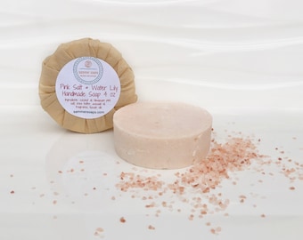 Pode incluir: Dois sabonetes artesanais de sal rosa e nenúfar. Um está desembrulhado, cilíndrico e rodeado por cristais de sal rosa. O outro está embrulhado em papel castanho com um rótulo que diz "Pink Salt + Water Lily Handmade Soap 4 oz."