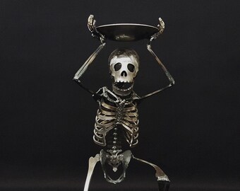 May include: A metal skeleton sculpture kneeling with its arms raised, holding a tray above its head. The sculpture is made of dark steel and has a realistic skull.
