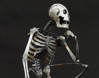 May include: A metal sculpture of a skeleton sitting on a sphere, with its hand on its chin, in a thinking pose. The sculpture is made of dark steel and is shown against a black background. "SHOWN IN DARK STEEL" is written below the sculpture.