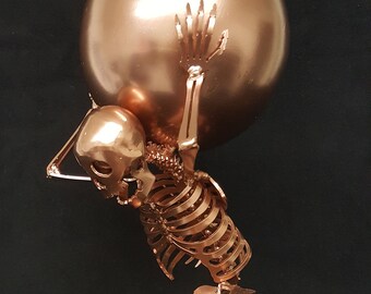May include: A copper-coloured metal sculpture of a skeleton holding a large sphere above its head. The sculpture is on a square base.
