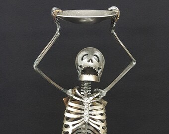 May include: A metal skeleton sculpture holding a bowl in its hands. The sculpture is made of recycled metal and has a dark steel finish. The sculpture is standing on a round base. "SHOWN IN DARK STEEL" is written below the sculpture.