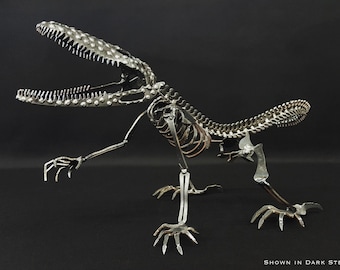 May include: A metal dinosaur skeleton sculpture with a large open mouth, made of small metal pieces and rivets. The sculpture is posed in a standing position with its tail curled up. The sculpture is shown in dark steel.