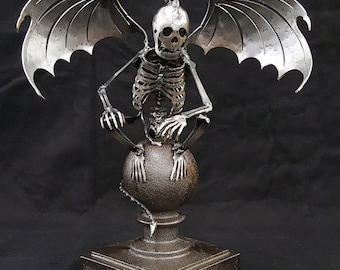 May include: A dark steel sculpture of a skeleton with large, stylized wings. The skeleton is perched atop a sphere, mounted on a square base. The sculpture has a detailed rib cage and skull. The text "SHOWN IN DARK STEEL" is at the bottom.