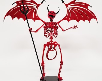 May include: Red metal skeleton sculpture of a devil with black wings and a trident. The sculpture is standing on a black base.