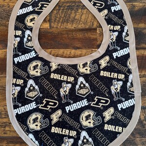 May include: A black baby bib with a tan trim and two snap closures. The bib is patterned with the words "PURDUE" and "BOILER UP", along with images of a football helmet and a mascot.
