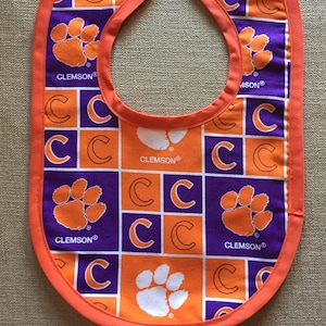 May include: Orange and purple baby bib with a Clemson University logo pattern. The bib has a white paw print and the text "CLEMSON®" repeated throughout the pattern.