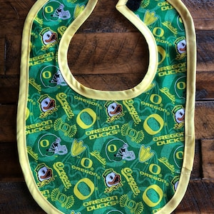 May include: A green and yellow baby bib with a repeating pattern of the Oregon Ducks logo, a duck mascot, and the words "Oregon Ducks".