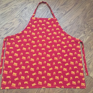 May include: Red apron with a repeating pattern of the USC Trojans logo in yellow. The apron has two pockets and adjustable ties.