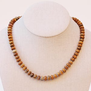 May include: A beaded necklace with natural-toned wooden beads. The beads are a mix of light and dark brown, strung together to form a necklace. The necklace is displayed on a white mannequin.
