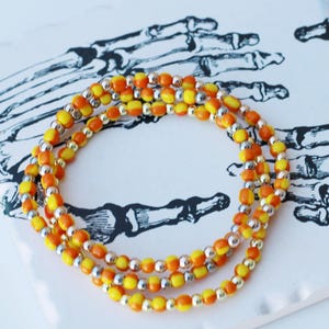 Può includere: Una collana di perline arancioni, gialle e argentate è drappeggiata su un sottobicchiere bianco. Il sottobicchiere presenta un'illustrazione in bianco e nero di una mano di scheletro. Un accessorio vivace.