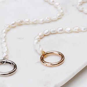 May include: Two necklaces featuring irregular, white pearls. One has a silver clasp, the other a gold clasp. They are presented on a white marble surface.