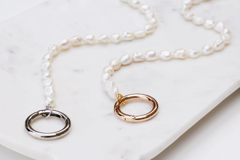 May include: Two necklaces featuring irregular, white pearls. One has a silver clasp, the other a gold clasp. They are presented on a white marble surface.