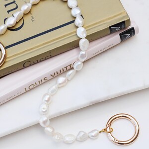 May include: A white pearl necklace with gold clasps and connectors. The necklace is draped over two books, one titled "Gucci" and the other "Louis Vuitton". The books are on a white surface.