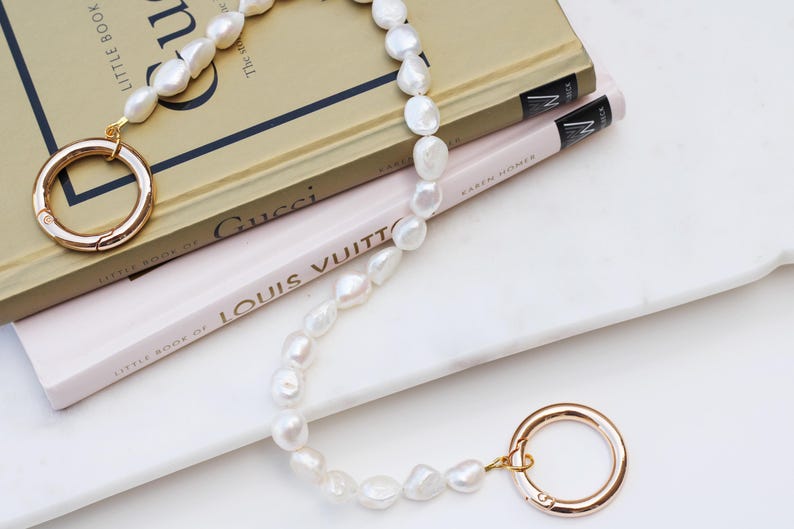 May include: A white pearl necklace with gold clasps and connectors. The necklace is draped over two books, one titled "Gucci" and the other "Louis Vuitton". The books are on a white surface.