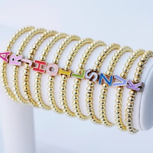 Op de afbeelding: Een set gouden kralen armbanden met kleurrijke letter bedels die de naam "HOLIDAY" spellen.