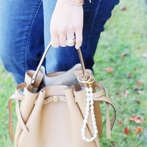May include: A tan leather bucket bag with a gold clasp and a pearl keychain. The bag is held by a person wearing blue jeans and a sweater. A collection of bracelets adorns the wrist.