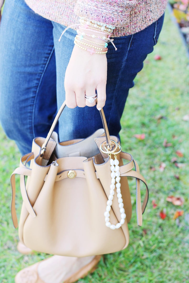May include: A tan leather bucket bag with a gold clasp and a pearl keychain. The bag is held by a person wearing blue jeans and a sweater. A collection of bracelets adorns the wrist.