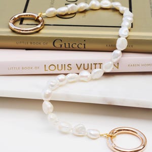May include: A pearl necklace with gold-coloured clasps lies on a stack of books. The necklace features irregular, white pearls and two large, circular clasps. The books are titled "Little Book of Gucci" and "Little Book of Louis Vuitton."