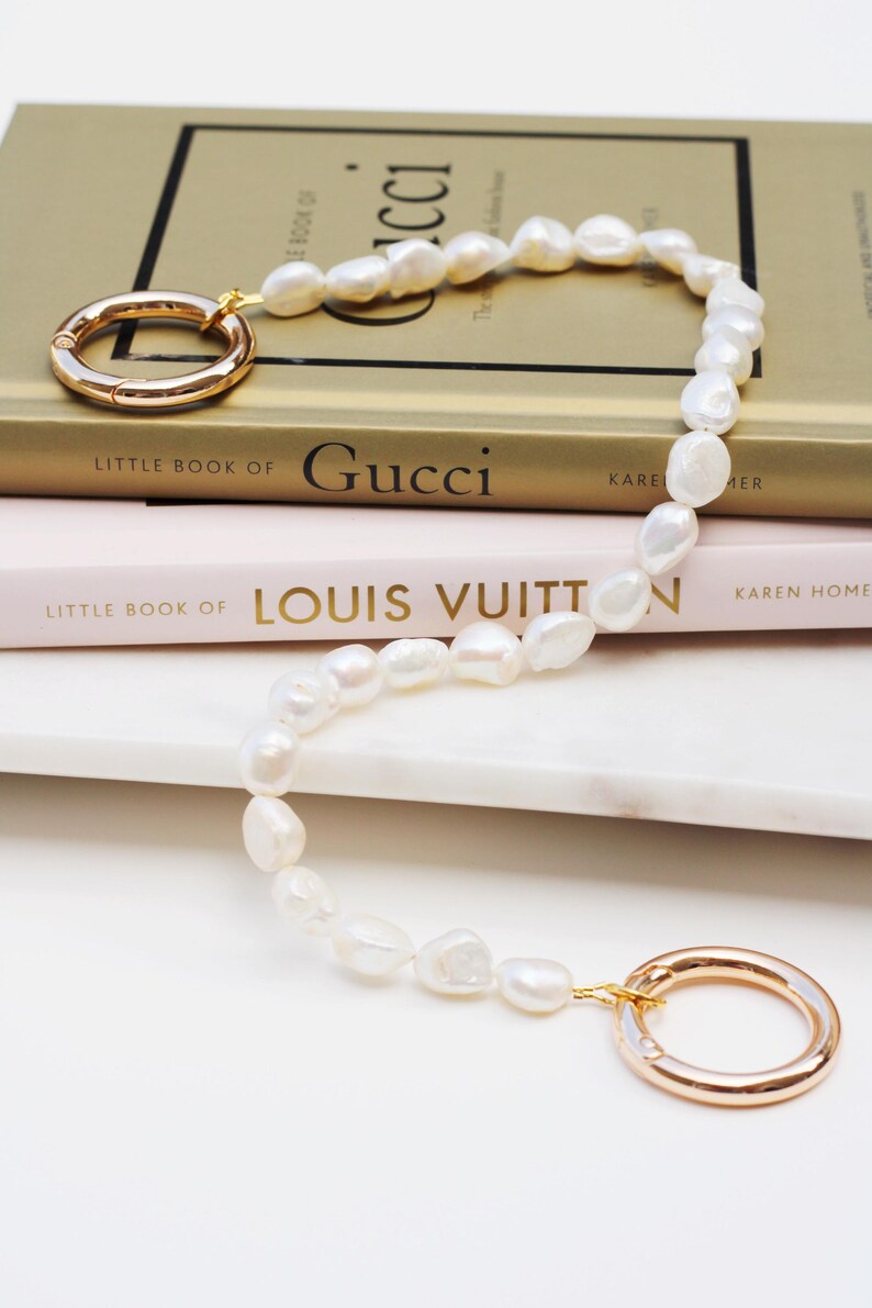 May include: A pearl necklace with gold-coloured clasps lies on a stack of books. The necklace features irregular, white pearls and two large, circular clasps. The books are titled "Little Book of Gucci" and "Little Book of Louis Vuitton."