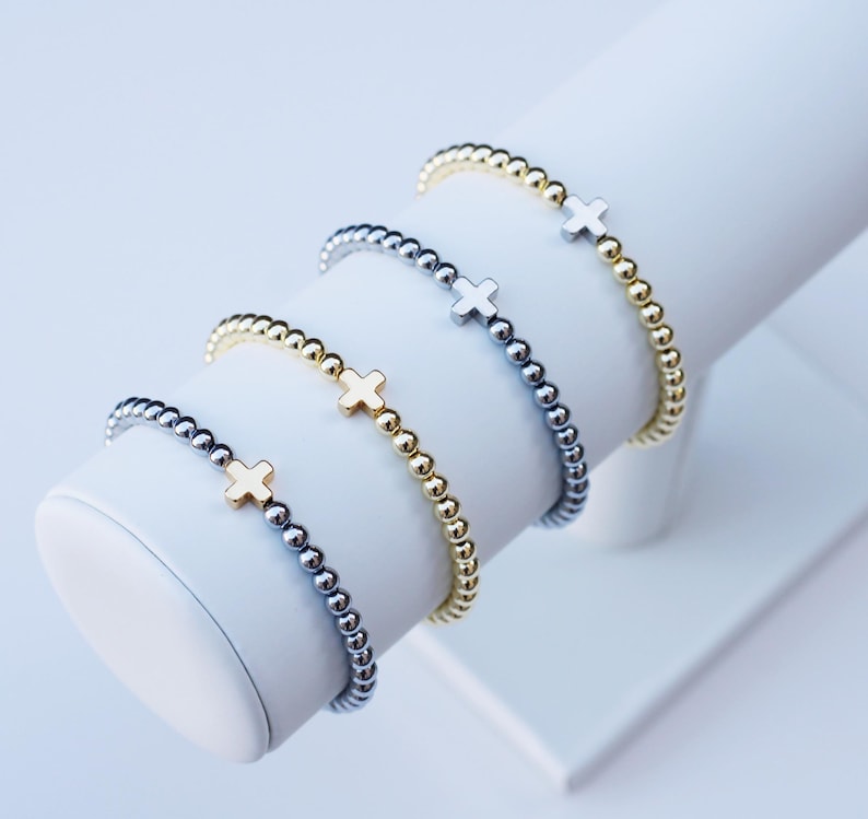 May include: Four beaded bracelets arranged in a row, each featuring a small cross charm. The bracelets alternate between gold-toned and silver-toned metallic beads, with matching metallic cross charms centered on each individual bracelet strand.