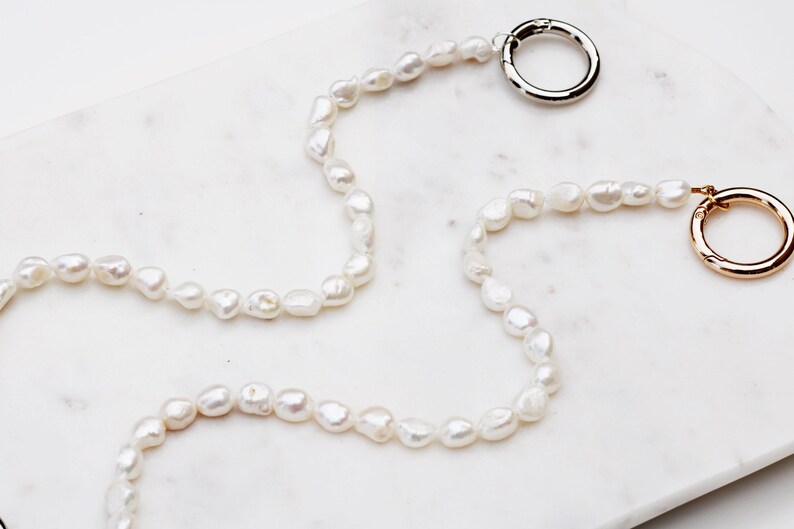 May include: A necklace made of irregular, white freshwater pearls. It has a silver-coloured clasp at one end and a gold-coloured clasp at the other. The necklace is displayed on a white marble surface.