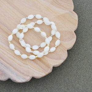 May include: Three white beaded bracelets with gold, silver, and rose gold accents are arranged on a light brown, scalloped-edge wooden dish. The beads are oval-shaped and strung together.