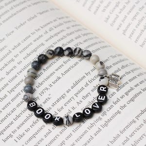 Puede incluir: Una pulsera de cuentas veteadas en blanco y negro y cuentas de letras negras que deletrean "BOOK LOVER". Un pequeño dije plateado de una pila de libros cuelga de la pulsera. La pulsera está sobre un libro abierto.