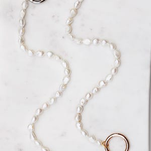 May include: A pearl necklace with three metal clasps. Two clasps are silver, and one is gold. The pearls are white and irregularly shaped. The necklace is displayed on a white marble surface.