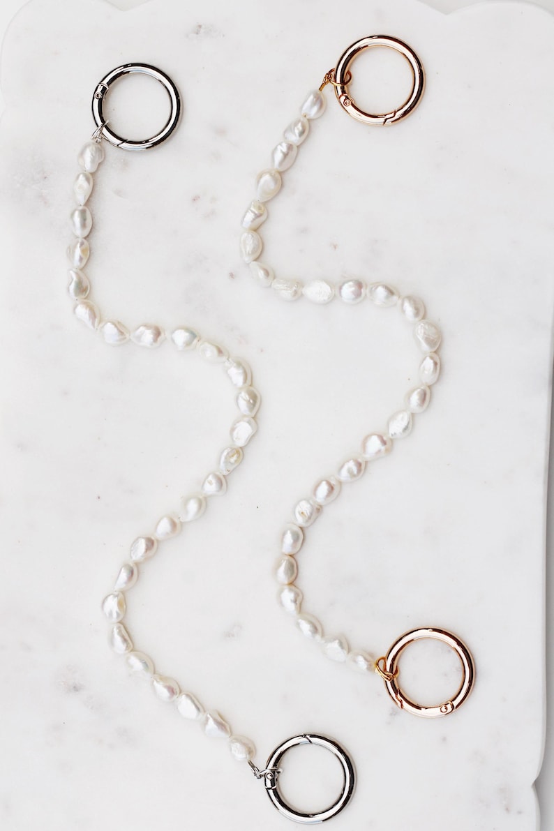 May include: A pearl necklace with three metal clasps. Two clasps are silver, and one is gold. The pearls are white and irregularly shaped. The necklace is displayed on a white marble surface.