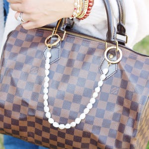 May include: A brown and tan checked handbag with a pearl chain. The bag features gold-tone hardware, a zip closure, and a padlock. The pearl chain is attached to the handles with gold rings.