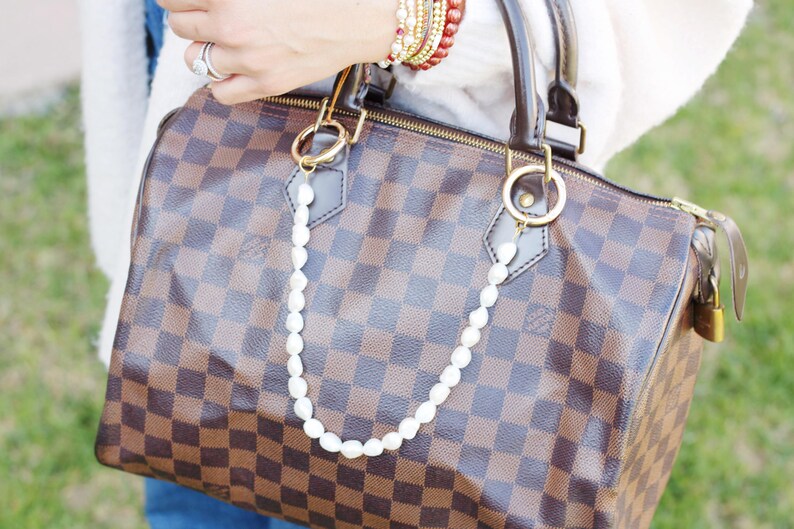 May include: A brown and tan checked handbag with a pearl chain. The bag features gold-tone hardware, a zip closure, and a padlock. The pearl chain is attached to the handles with gold rings.