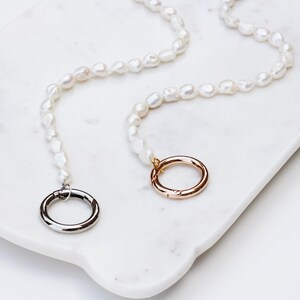 May include: Two pearl necklaces with irregular pearls and silver and gold clasps. The necklaces are displayed on a white marble tray. The clasps are circular. The pearls are a creamy white colour, and the overall aesthetic is elegant.