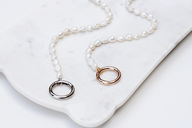 May include: Two pearl necklaces with irregular pearls and silver and gold clasps. The necklaces are displayed on a white marble tray. The clasps are circular. The pearls are a creamy white colour, and the overall aesthetic is elegant.