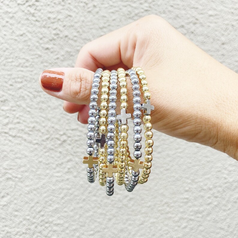 Puede incluir: Una pila de pulseras de cuentas doradas y plateadas con peque&ntilde;os dijes de cruz. Cada pulsera est&aacute; hecha de cuentas diminutas y tiene un delicado dije de cruz.