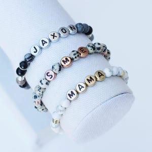 Puede incluir: Tres pulseras de cuentas con cuentas con letras que deletrean "JAXON", "SME" y "MAMA". Las pulseras presentan una mezcla de cuentas negras, grises, blancas y doradas. Las pulseras se exhiben sobre una superficie blanca texturizada.
