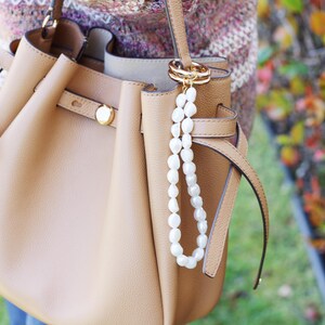 May include: A tan leather handbag with a gold clasp and a white pearl beaded charm. The bag has a structured design with a top handle and a shoulder strap. The background is blurred, with hints of green and autumn colours.