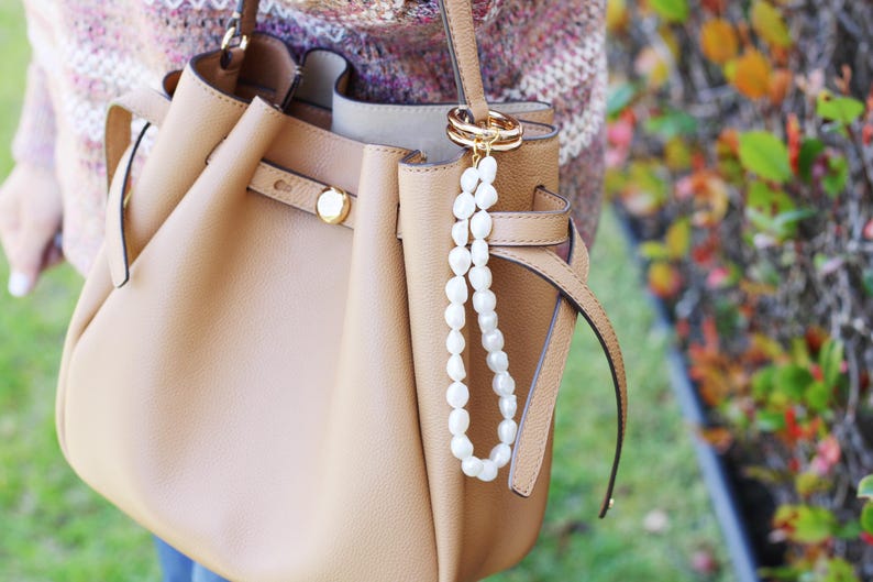 May include: A tan leather handbag with a gold clasp and a white pearl beaded charm. The bag has a structured design with a top handle and a shoulder strap. The background is blurred, with hints of green and autumn colours.