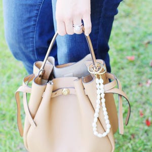 May include: A tan leather handbag with a drawstring closure and gold-tone hardware. A pearl beaded charm hangs from the handle. The bag is held by a person wearing blue jeans.