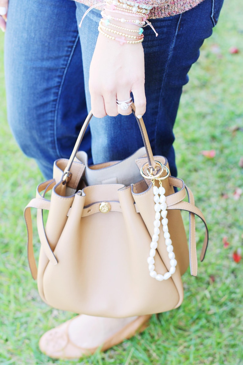 May include: A tan leather handbag with a drawstring closure and gold-tone hardware. A pearl beaded charm hangs from the handle. The bag is held by a person wearing blue jeans.