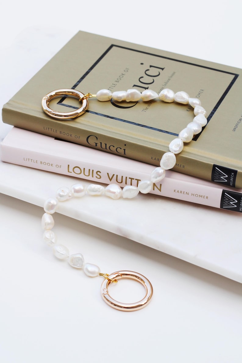 May include: A white pearl necklace with gold-coloured circular clasps. The necklace is draped over a stack of books, including "Little Book of Gucci" and "Little Book of Louis Vuitton."