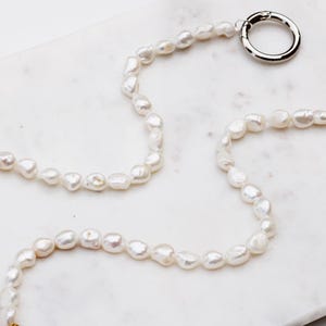 May include: A white pearl necklace with an irregular shape, featuring a silver and a gold clasp. The necklace is displayed on a white marble surface. The pearls are strung together.