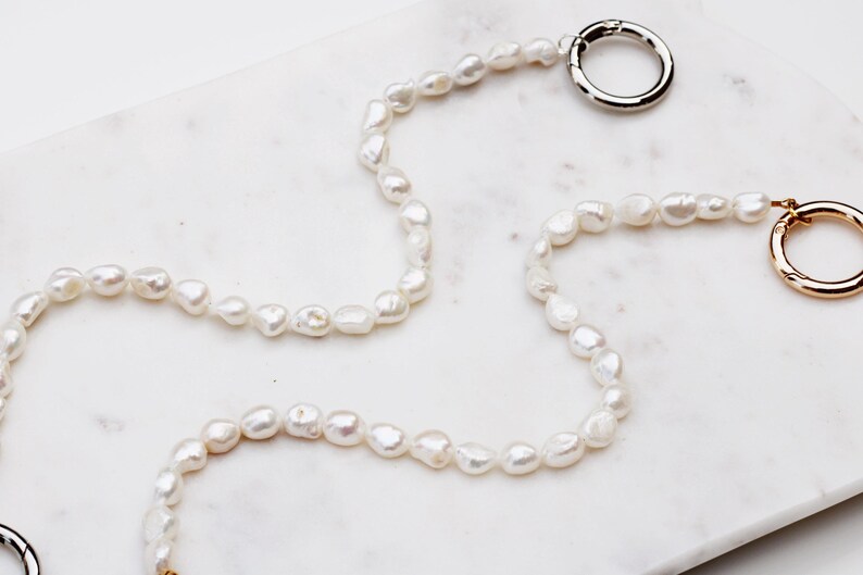 May include: A white pearl necklace with an irregular shape, featuring a silver and a gold clasp. The necklace is displayed on a white marble surface. The pearls are strung together.