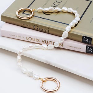 May include: A pearl necklace with gold-coloured circular clasps. The necklace is draped over a stack of books, including "Little Book of Gucci" and "Little Book of Louis Vuitton". The books are on a white surface.