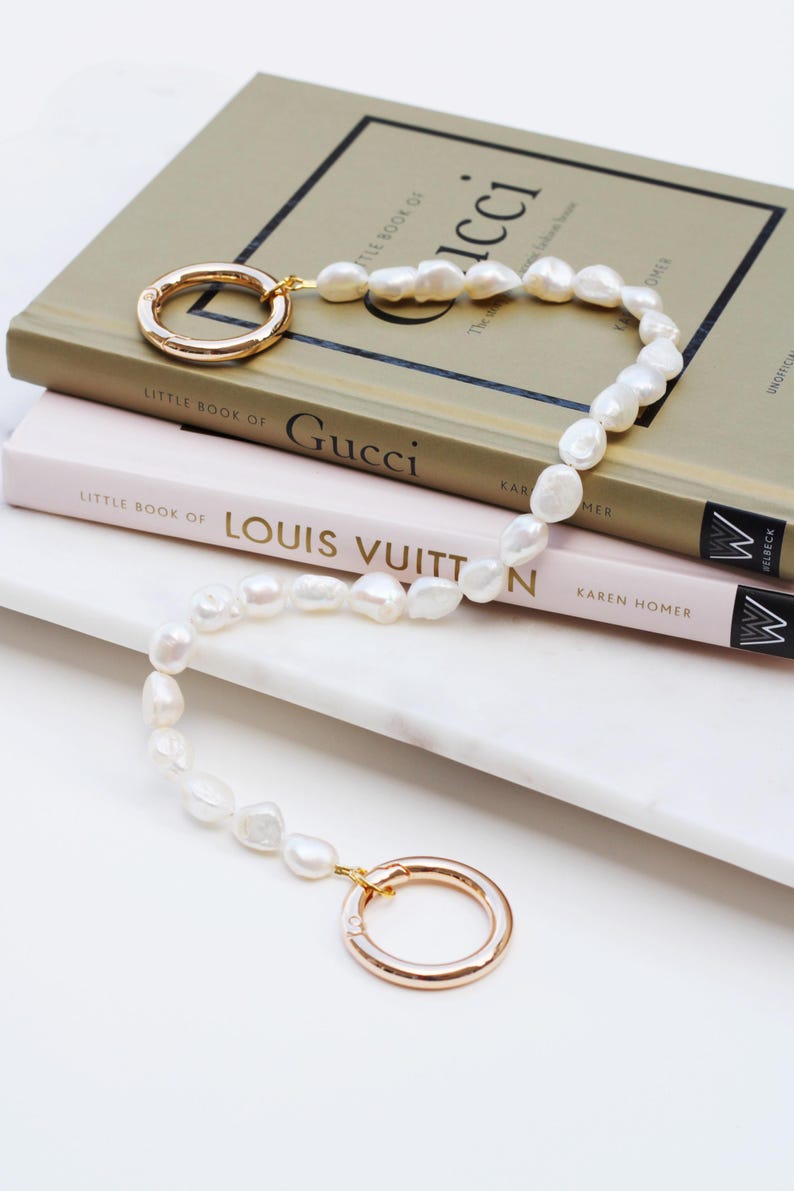 May include: A pearl necklace with gold-coloured circular clasps. The necklace is draped over a stack of books, including "Little Book of Gucci" and "Little Book of Louis Vuitton". The books are on a white surface.