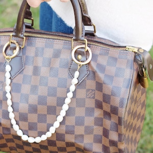 May include: A brown and tan checked handbag with gold-coloured zip and hardware. A string of white pearls hangs from the gold rings on the bag. The bag has brown leather handles and trim.
