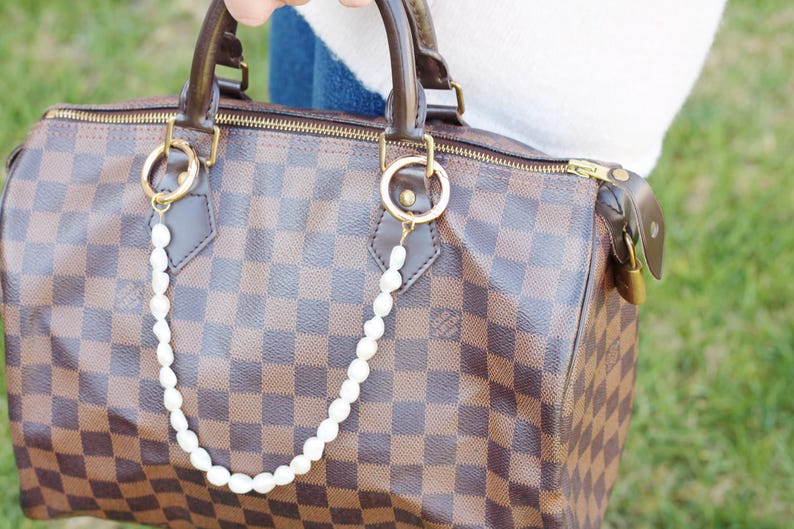 May include: A brown and tan checked handbag with gold-coloured zip and hardware. A string of white pearls hangs from the gold rings on the bag. The bag has brown leather handles and trim.