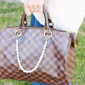 May include: A brown and tan checked handbag with a pearl chain attached to the gold-toned handles. The bag has a zip closure and a brown leather handle. The background is blurred green grass.