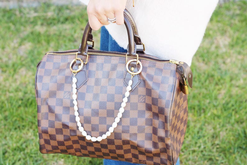 May include: A brown and tan checked handbag with a pearl chain attached to the gold-toned handles. The bag has a zip closure and a brown leather handle. The background is blurred green grass.